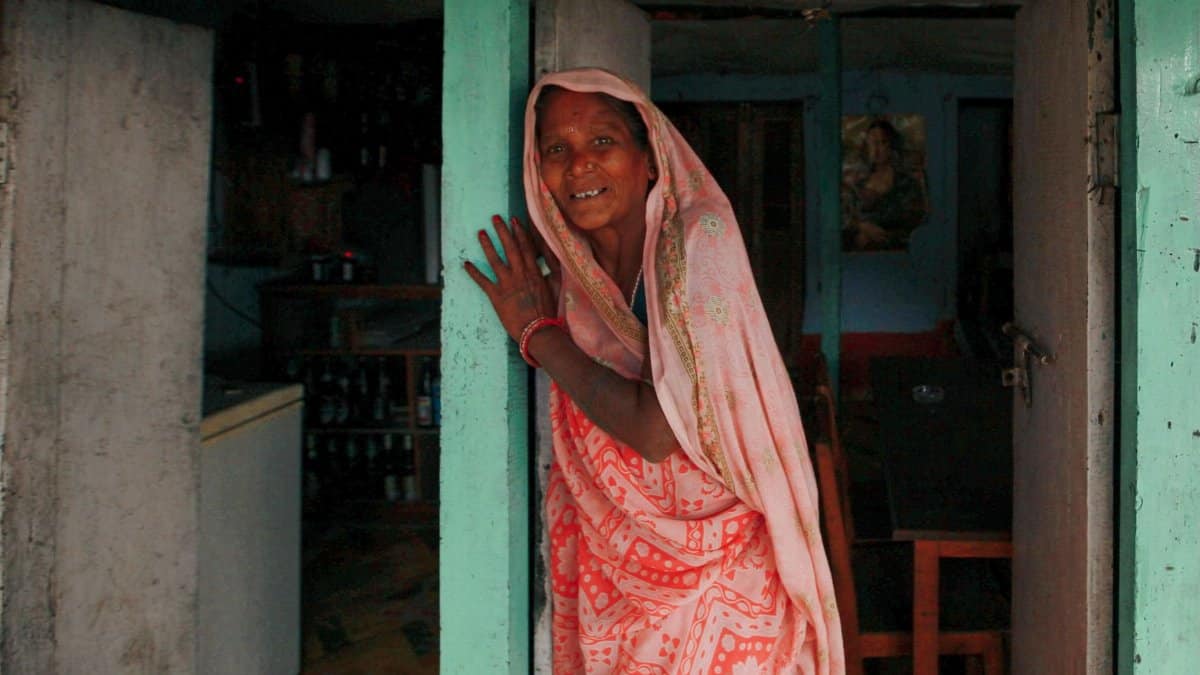 A middle-aged woman in a colorful sari smiles warmly while standing in a doorway.