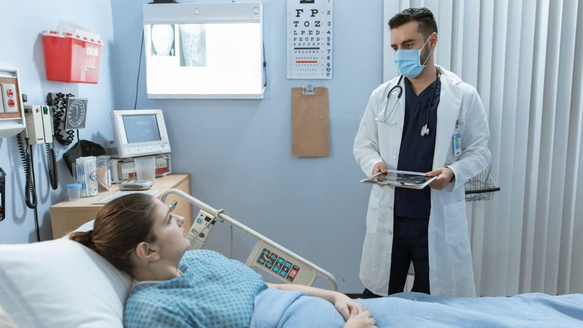 Doctor wearing face mask consulting with a patient in a hospital room, highlighting healthcare safety.