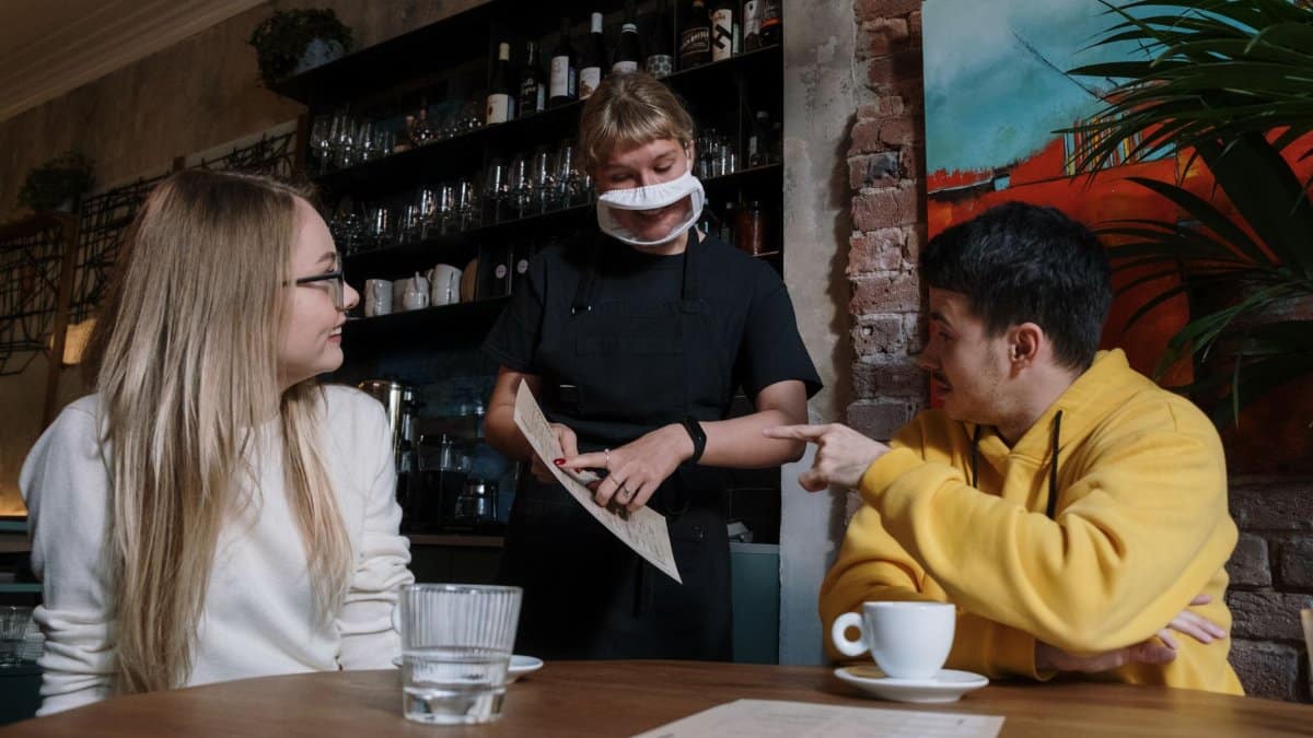 A masked waitress serves two patrons in a cozy restaurant setting.