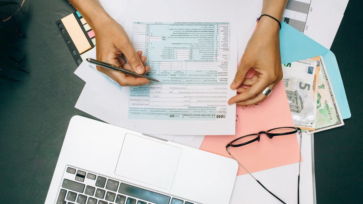 Hands writing on tax documents with laptop, glasses, and currency on desk.
