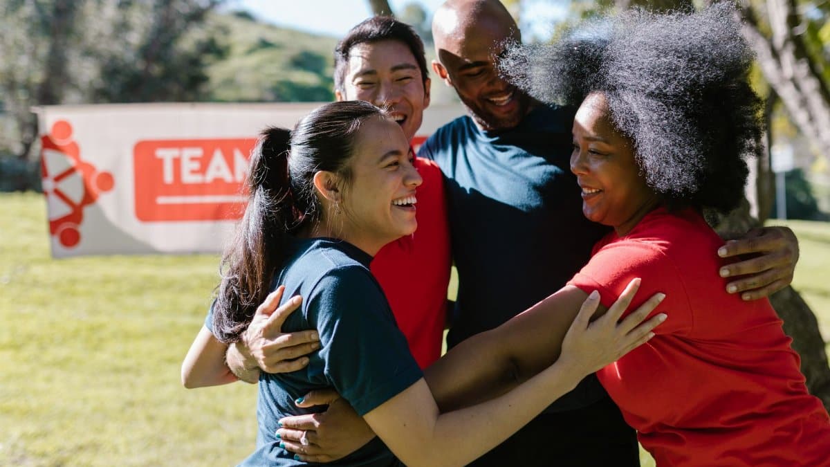 A joyful, diverse group embraces during an outdoor team building event in a sunny park.