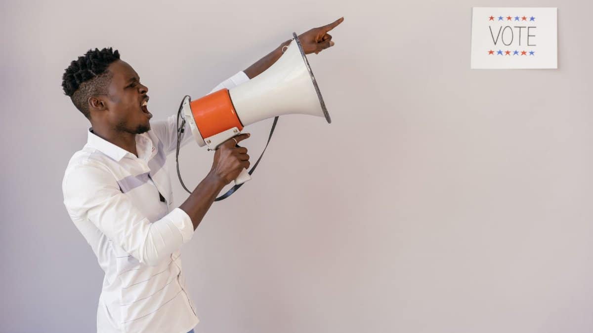 A passionate man urging people to vote using a megaphone, pointing at a sign.