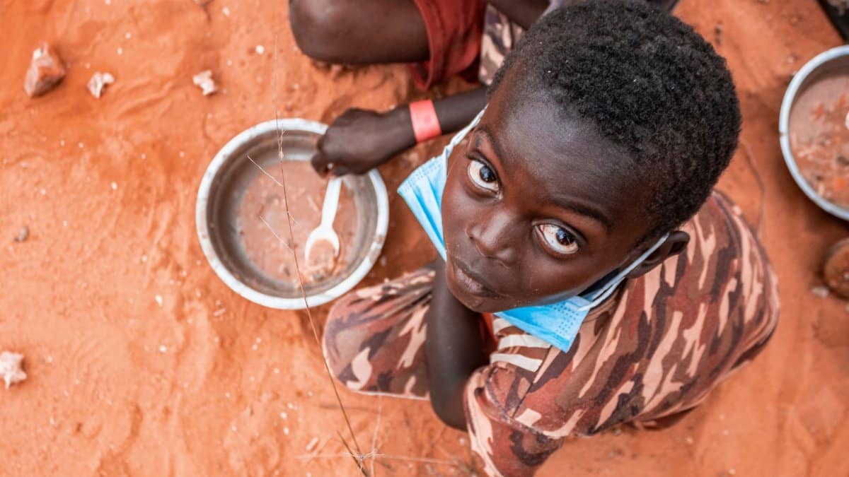 Child on a sandy ground in Luanda with mask and empty bowl, highlighting pandemic challenges.