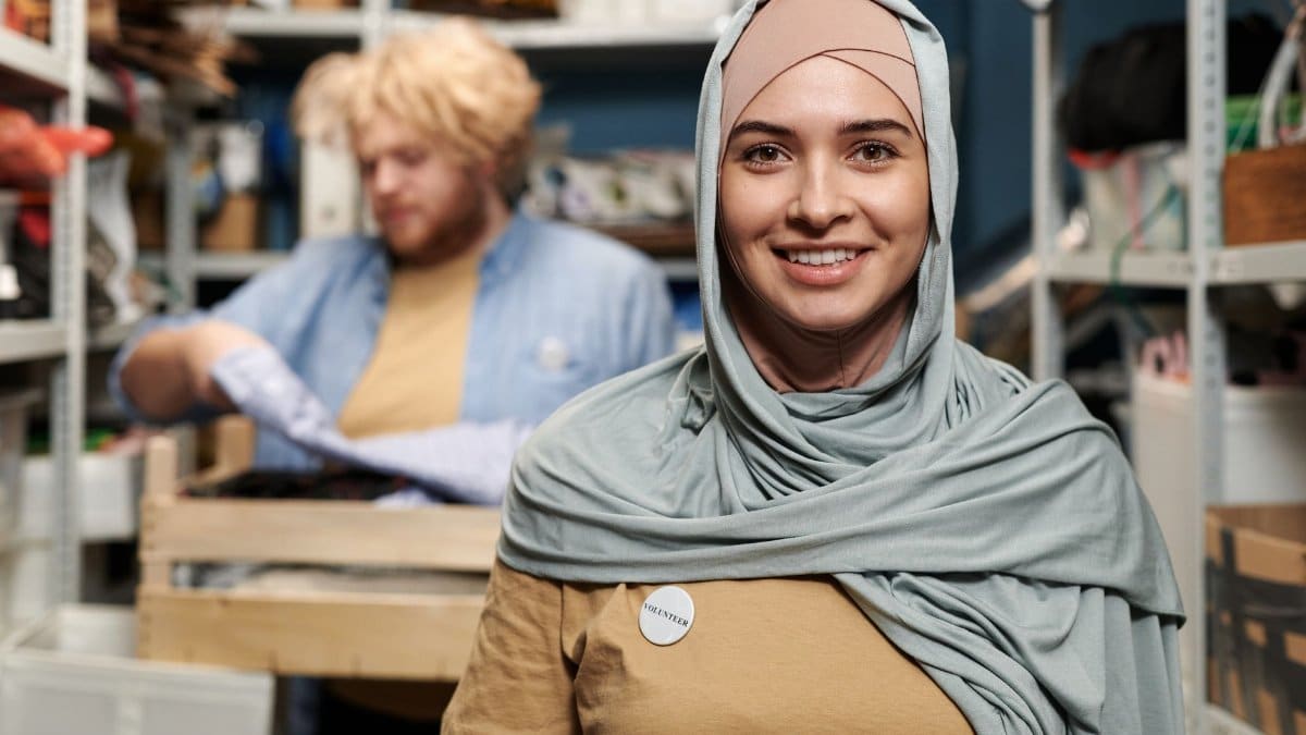 Smiling woman in hijab volunteering in a storeroom with a colleague organizing supplies.
