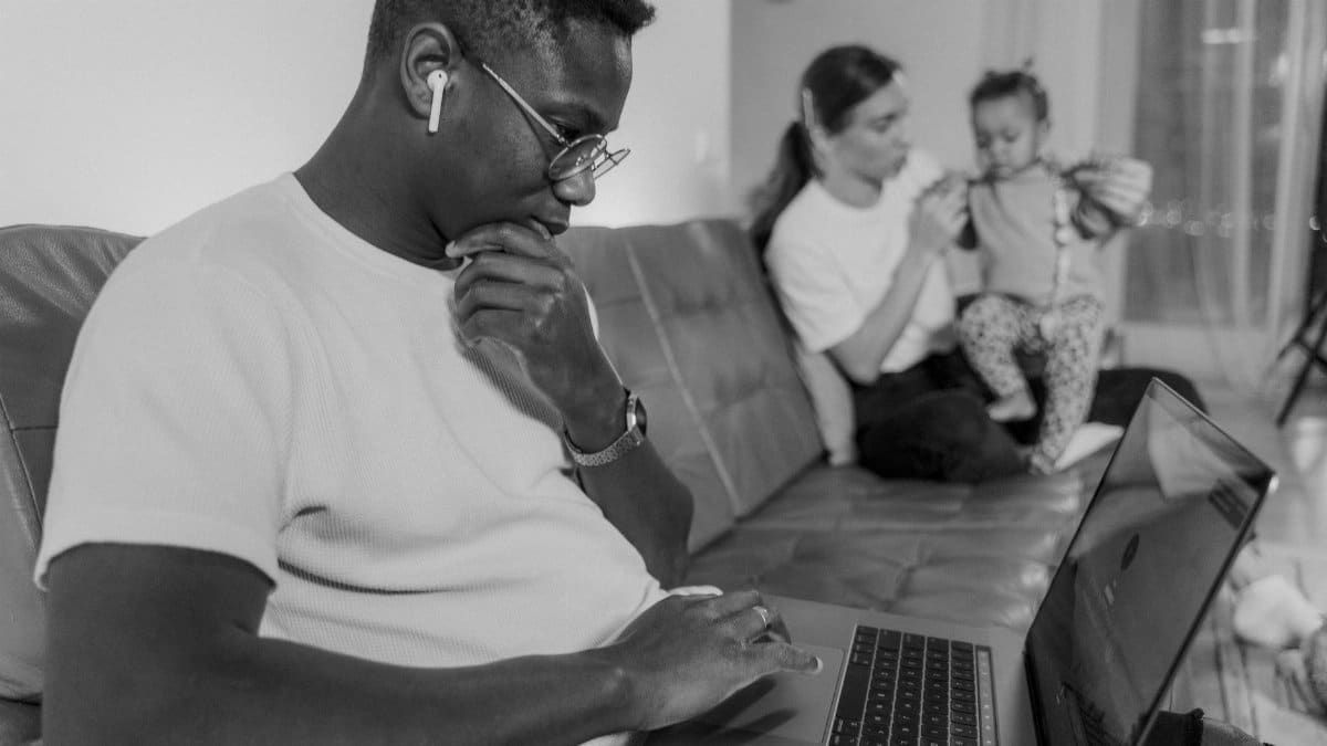 Black and white scene of a man working on a laptop while a woman cares for a child on a couch.