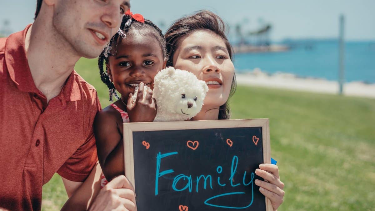 Happy mixed family bonding with a teddy bear and a chalkboard that reads 'Family' on a sunny day by the sea.