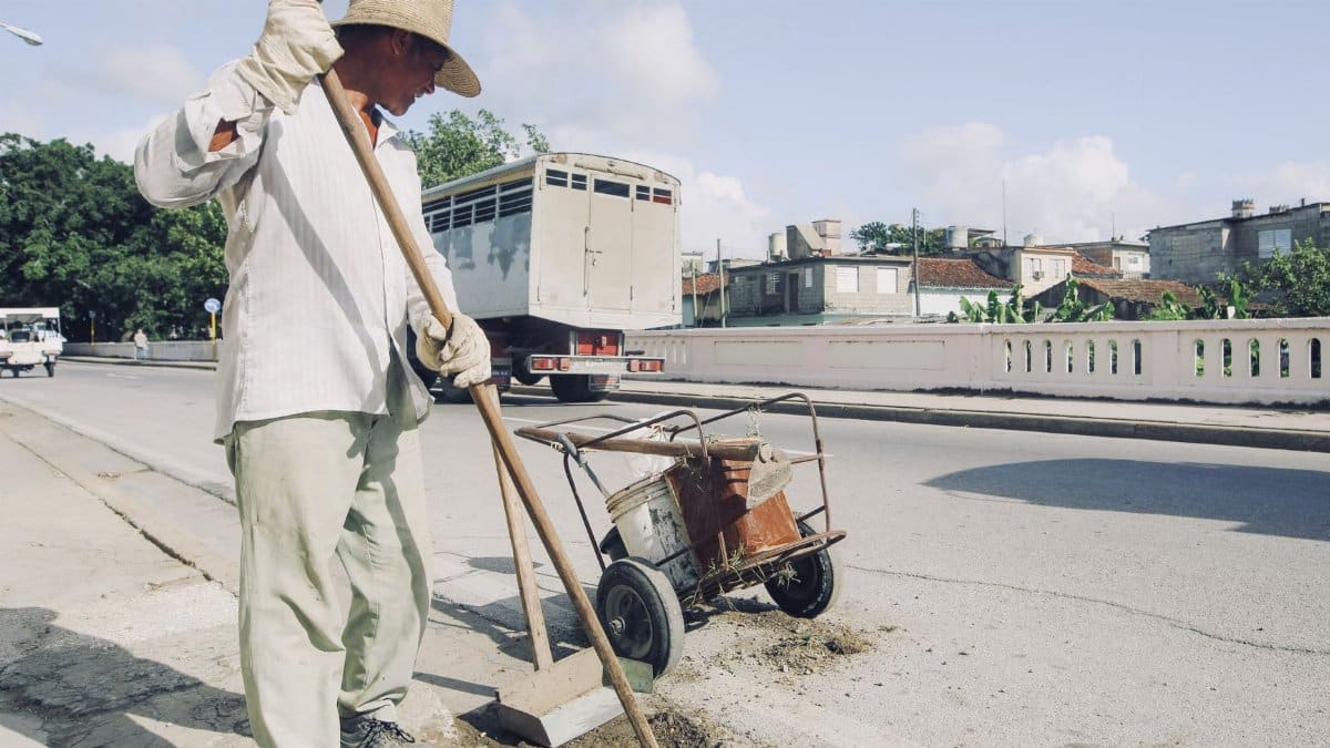 A city street cleaner in a hat sweeps the sidewalk on a sunny day with traffic in the background.