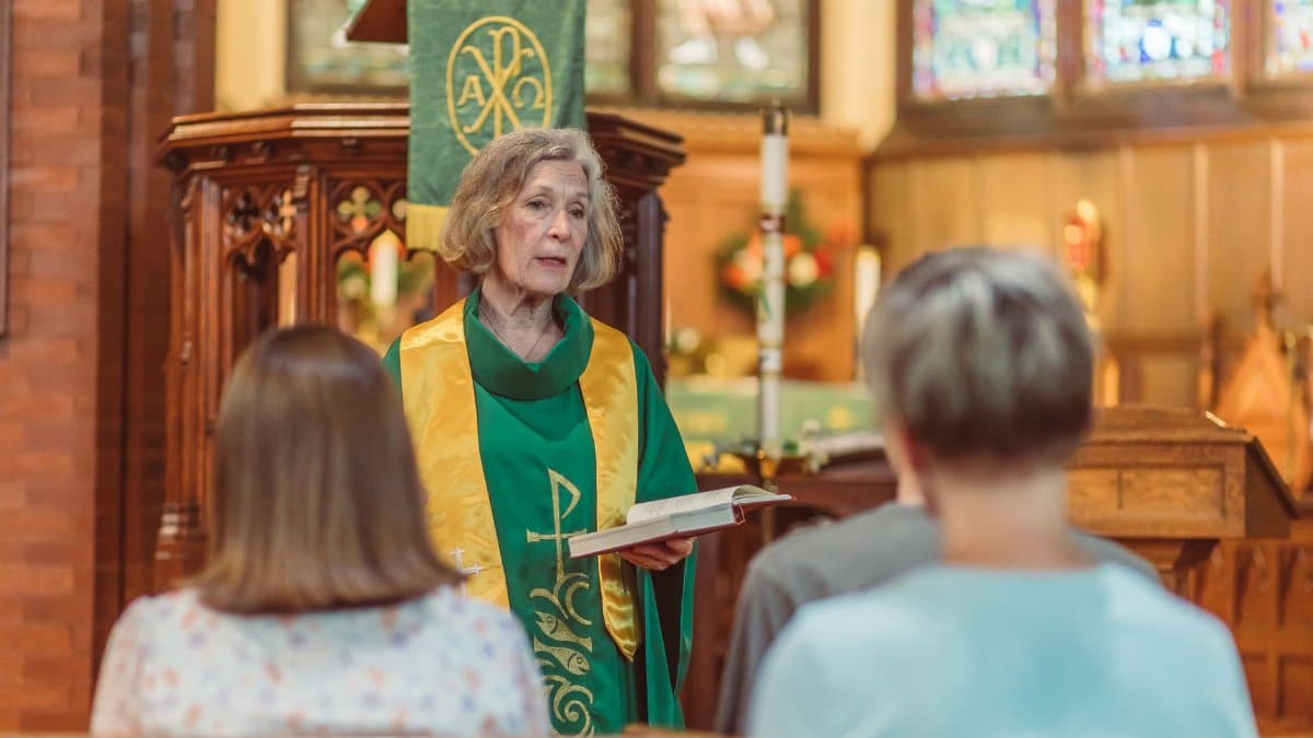An elderly female pastor delivers a sermon holding an open book in a church.