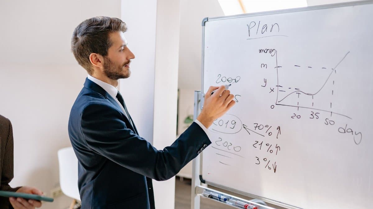 A businessman in a suit writes financial data on a whiteboard during an office planning session.