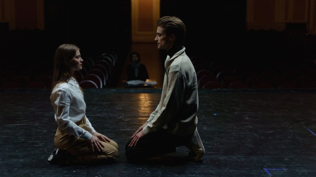 Two actors kneeling face-to-face in an emotional theater rehearsal, captured on a dimly lit stage.