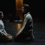 Two actors kneeling face-to-face in an emotional theater rehearsal, captured on a dimly lit stage.