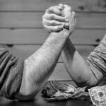 Two men engage in a high-stakes arm wrestling match surrounded by money on a table.