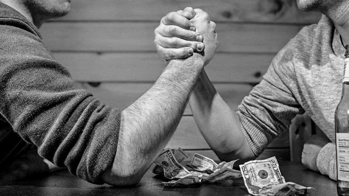 Two men engage in a high-stakes arm wrestling match surrounded by money on a table.