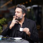 A contemplative middle-aged man enjoys coffee and a cigarette at an outdoor café.