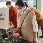 Voters casting ballots indoors on Election Day, wearing masks and using tablets for contactless voting.