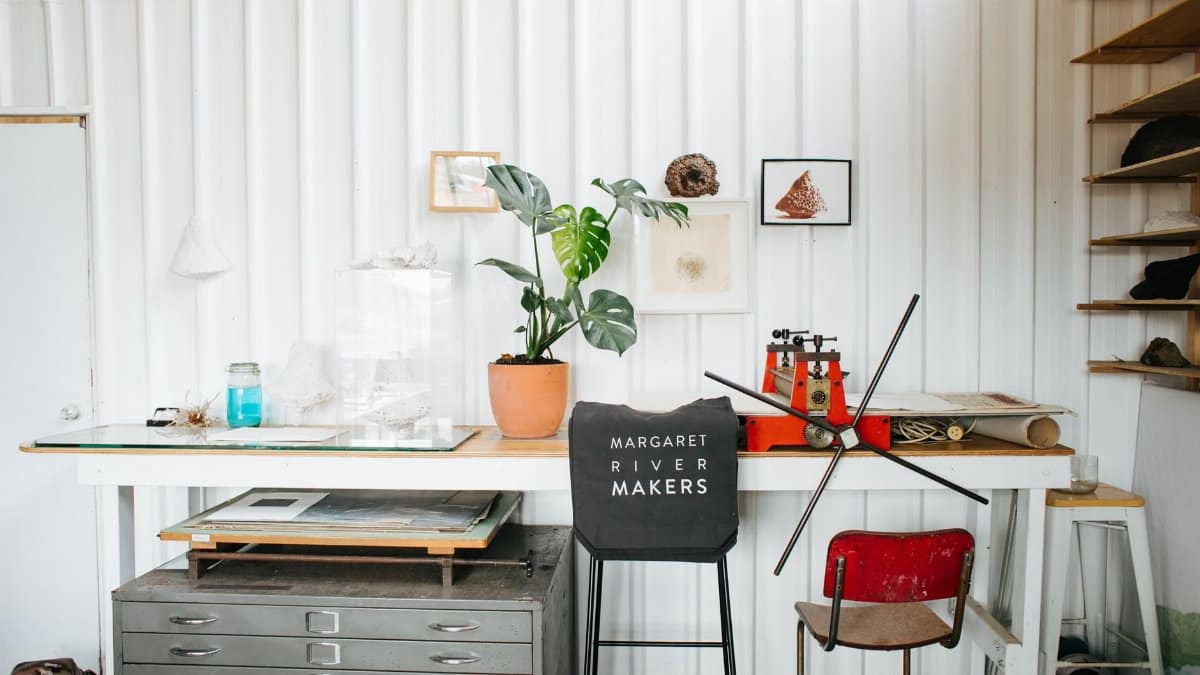 Interior of creative workplace with printing press and flowerpot placed on table near drawer and chair with manufactured product with inscription