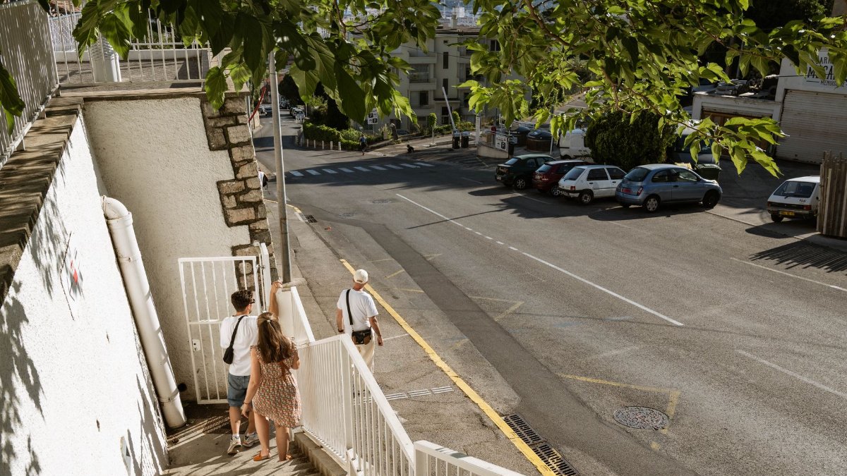 People walking down stairs toward a Toulon street on a sunny day, framed by green foliage.