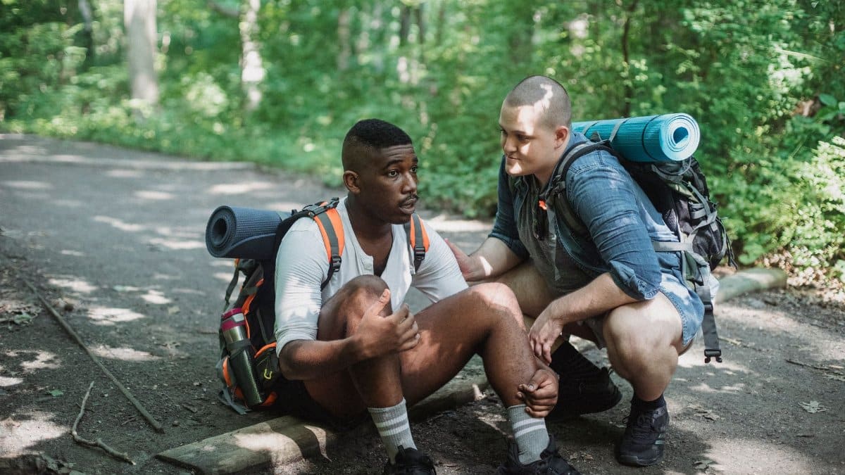 Full body of tired African American male traveler sitting on ground with friend during difficult trekking