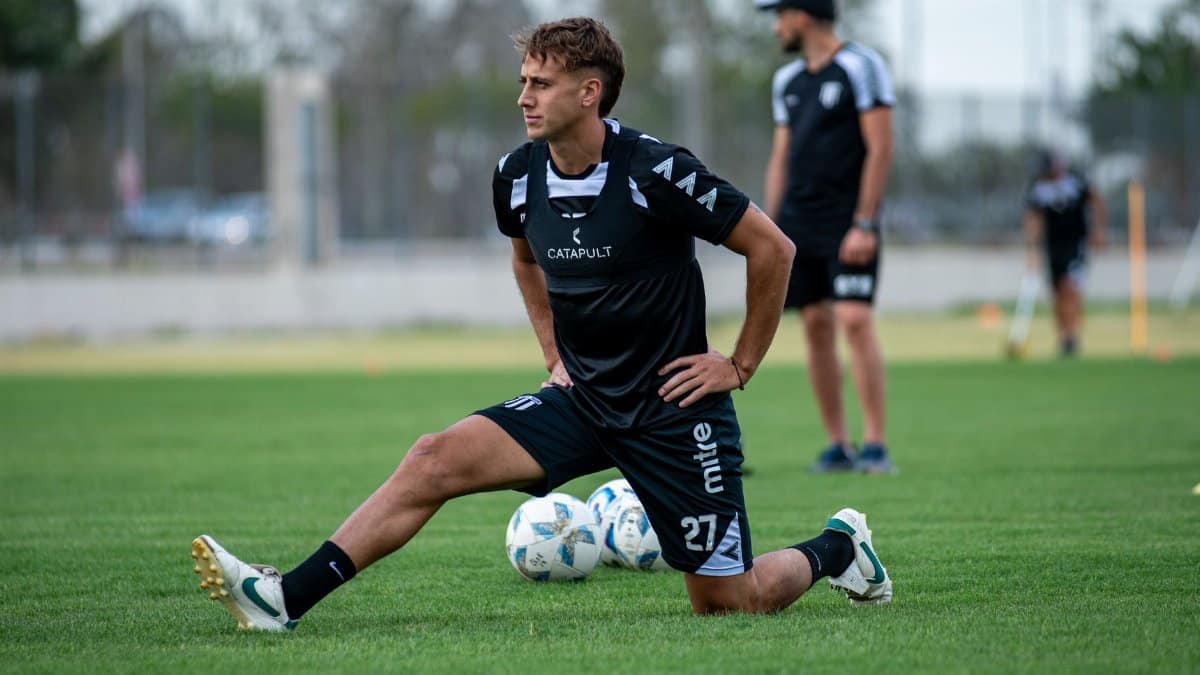 A soccer player stretches on the field during training, surrounded by teammates and soccer balls.