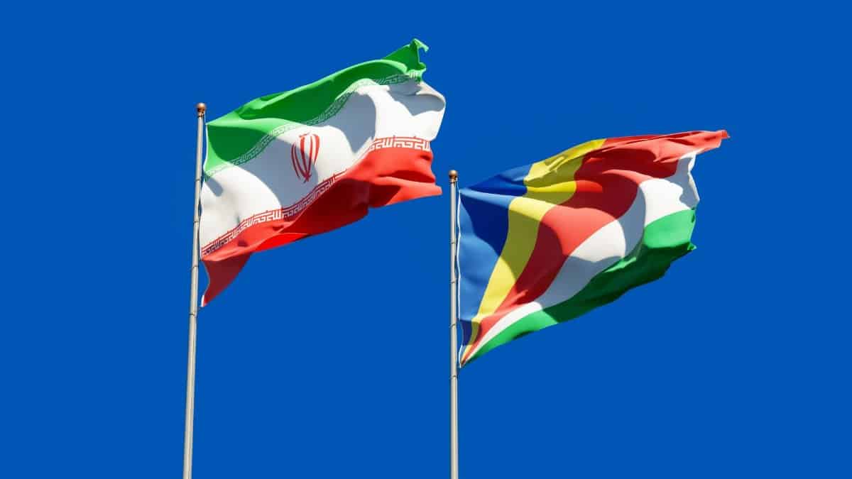 National flags of Iran and Seychelles waving on poles against a bright blue sky, symbolizing international relations.
