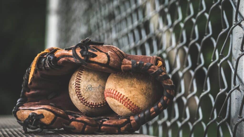 Close-up of a leather glove holding two vintage baseballs, resting near a chain-link fence.