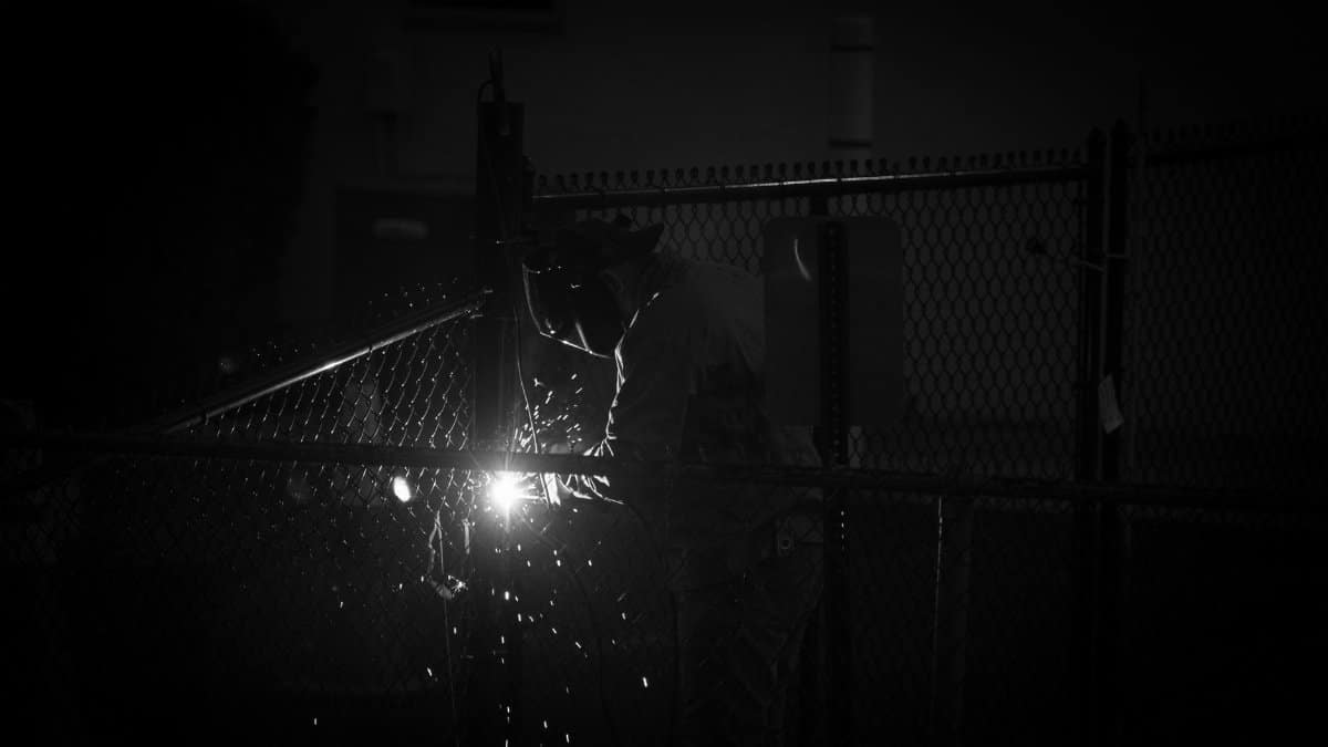 A welder works on a metal fence, sparks flying in a dramatic black and white scene.