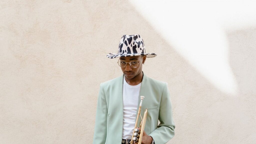 Fashionable musician wearing a fedora and holding a trumpet against a neutral background.