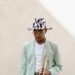 Fashionable musician wearing a fedora and holding a trumpet against a neutral background.