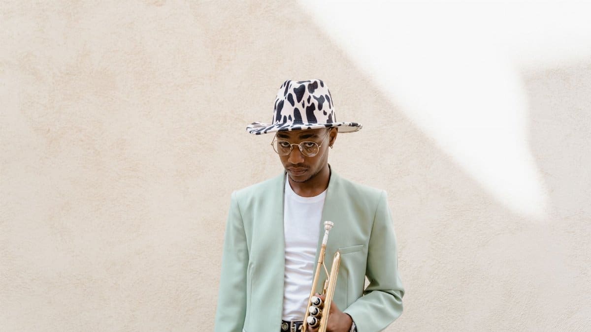 Fashionable musician wearing a fedora and holding a trumpet against a neutral background.