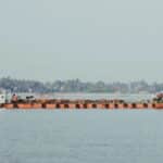 A cargo ship navigates a wide river near a lush, distant shoreline under a clear sky.
