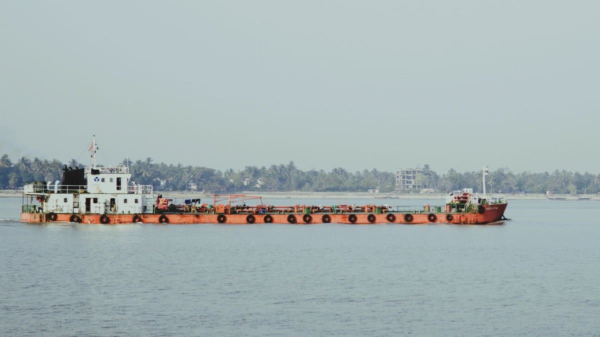 A cargo ship navigates a wide river near a lush, distant shoreline under a clear sky.