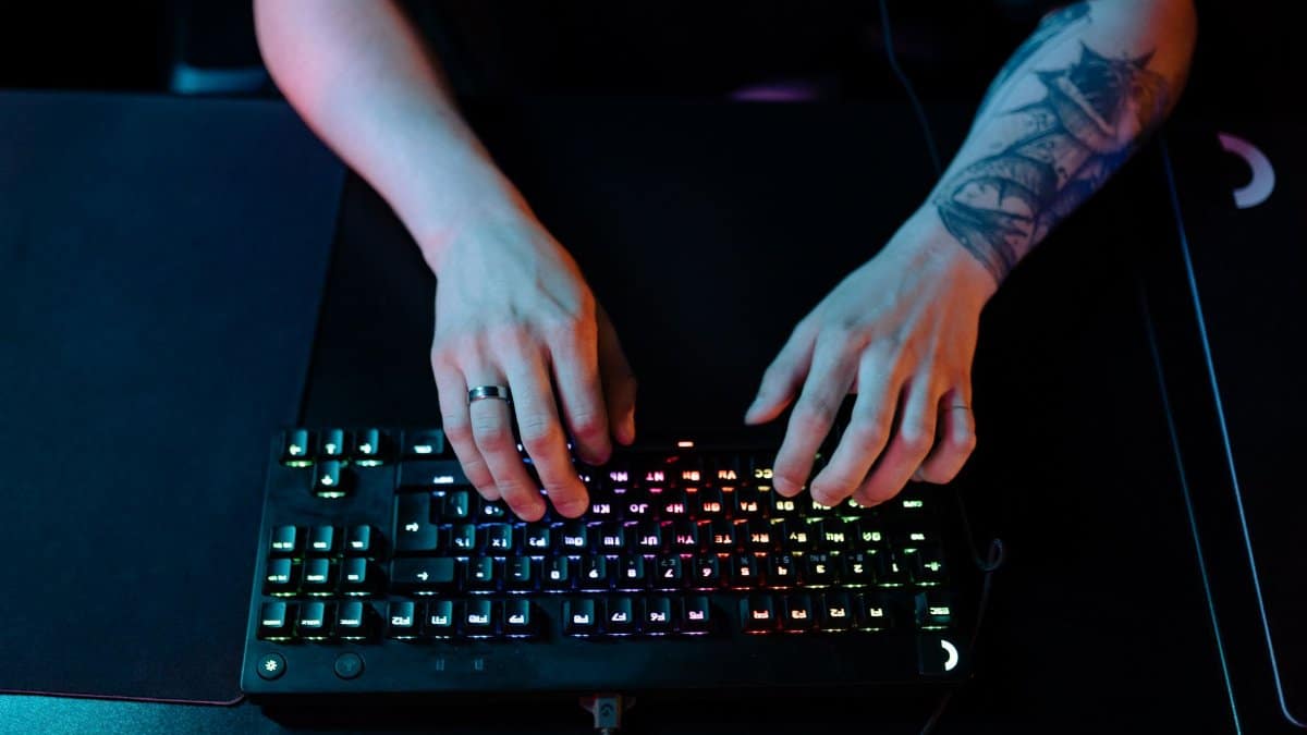 Close-up of hands typing on a RGB gaming keyboard with a tattooed arm and ring visible.