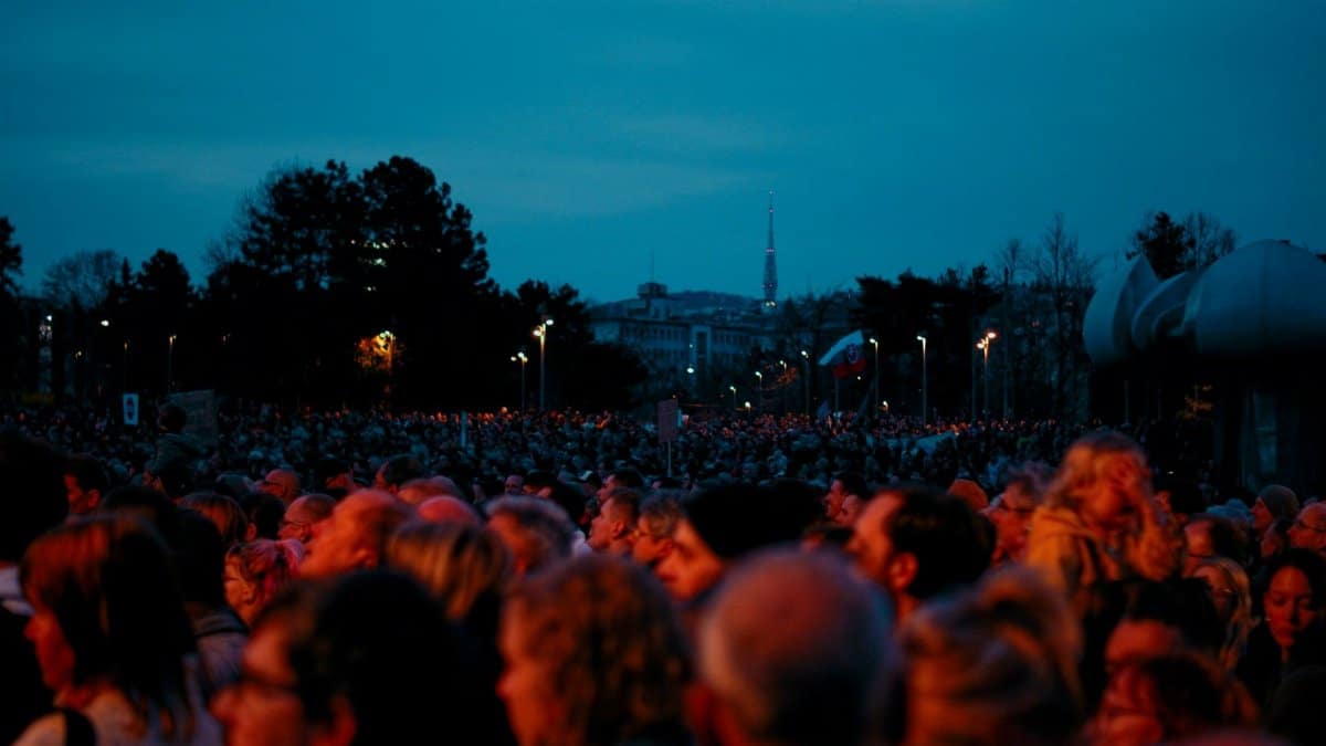 A large diverse crowd gathers outdoors at dusk for a public event, cityscape backdrop.