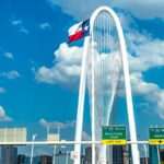 Iconic view of Margaret Hunt Hill Bridge with Texas flag and Dallas skyline under a blue sky.