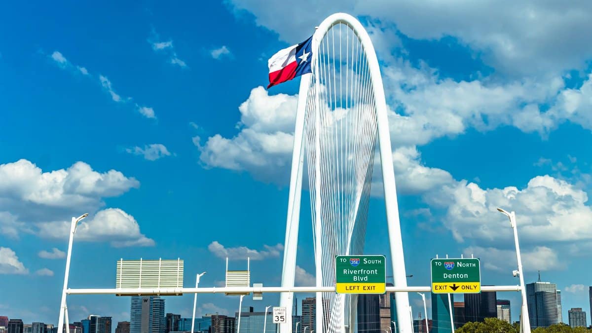 Iconic view of Margaret Hunt Hill Bridge with Texas flag and Dallas skyline under a blue sky.