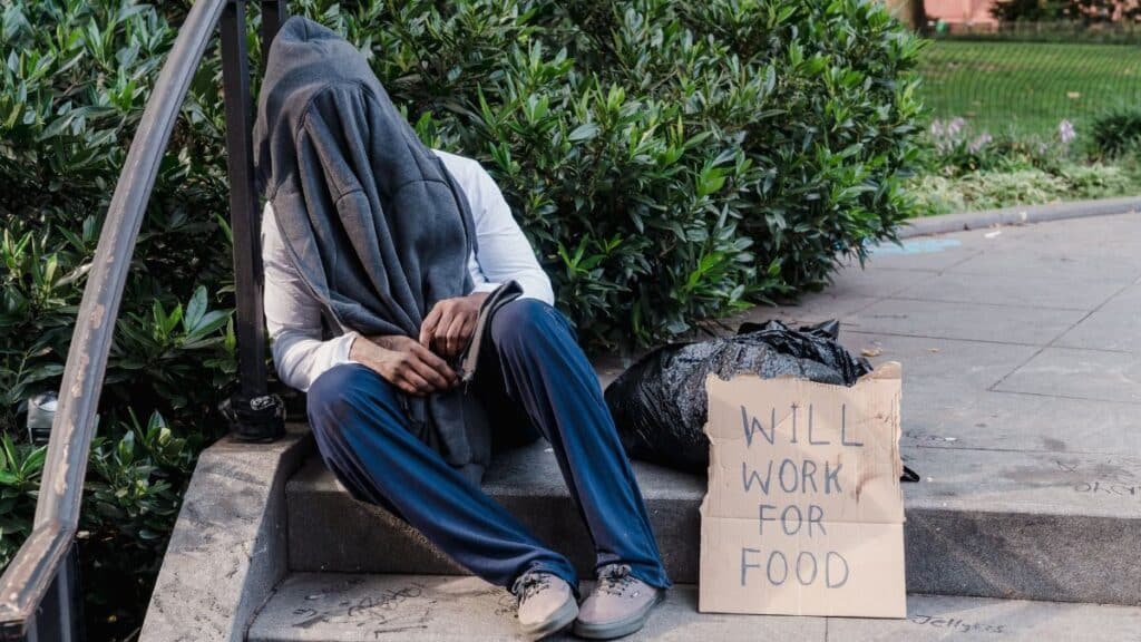 A homeless person sitting on outdoor stairs, covered by a cloth, with a sign reading 'Will Work for Food'.