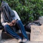 A homeless person sitting on outdoor stairs, covered by a cloth, with a sign reading 'Will Work for Food'.