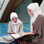 Two women wearing hijabs reading the Quran together inside a contemporary mosque with stylish design.