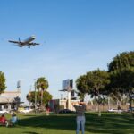 A passenger plane approaches for landing, flying low over a park with people watching.