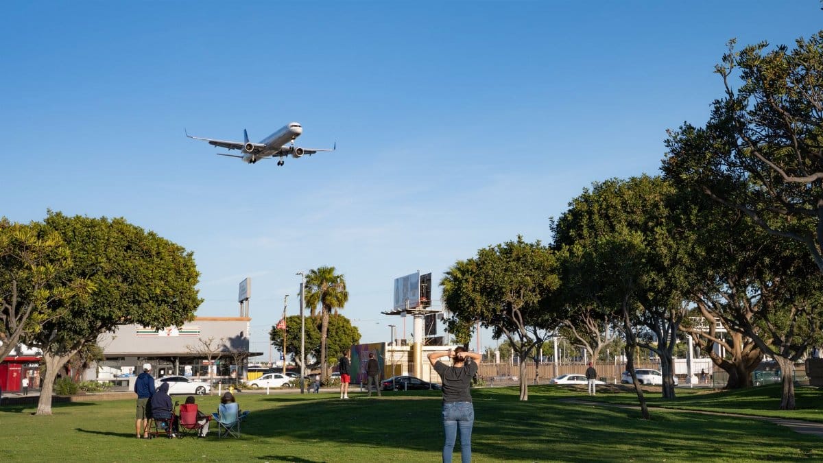 A passenger plane approaches for landing, flying low over a park with people watching.
