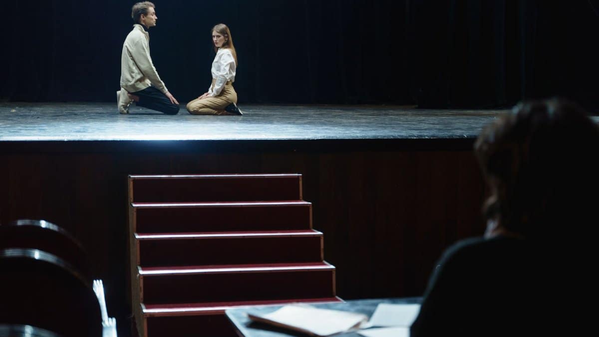 Actors in a dramatic rehearsal on stage, under dim theater lights.