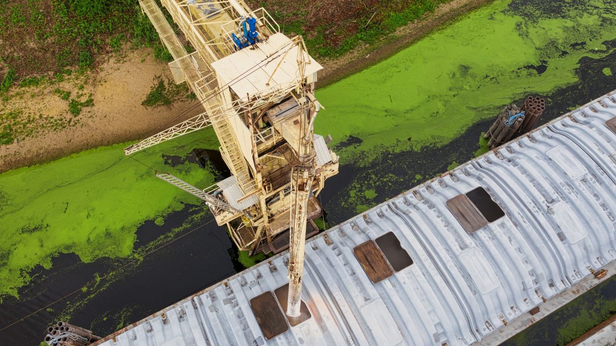 Aerial shot capturing an industrial crane over a river with green algae, highlighting industrial impact on nature.