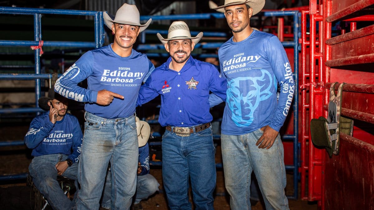 Three smiling cowboys pose at a rodeo in Tiros, Minas Gerais, Brazil, wearing cowboy hats and blue shirts.
