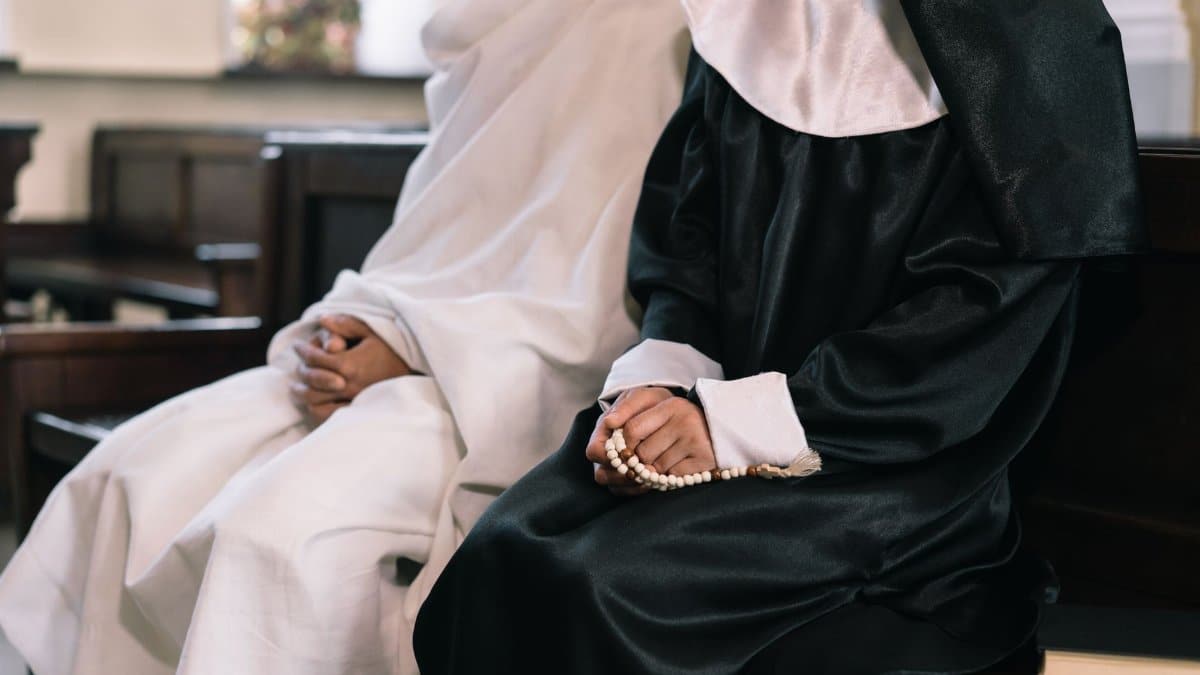 Two nuns in religious habits sitting and praying with rosary beads in a church setting.