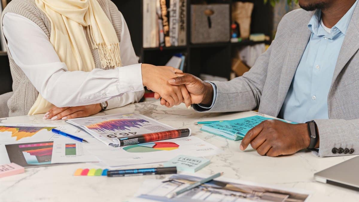 Two business professionals shaking hands over financial documents in an office setting.
