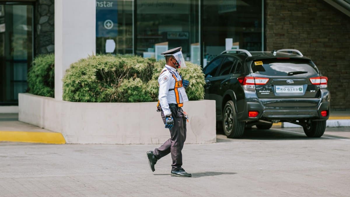 A security guard wearing a mask and gloves walks by a parked car outdoors, ensuring safety.