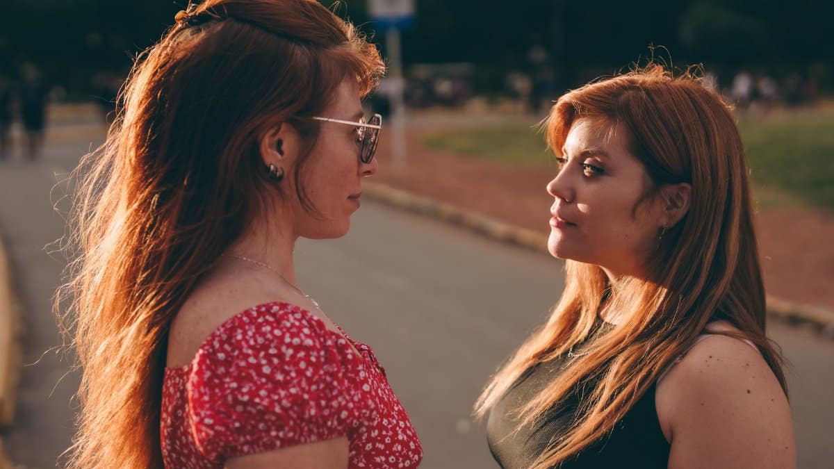 Two women with red hair standing outdoors, facing each other with sunlight highlights.