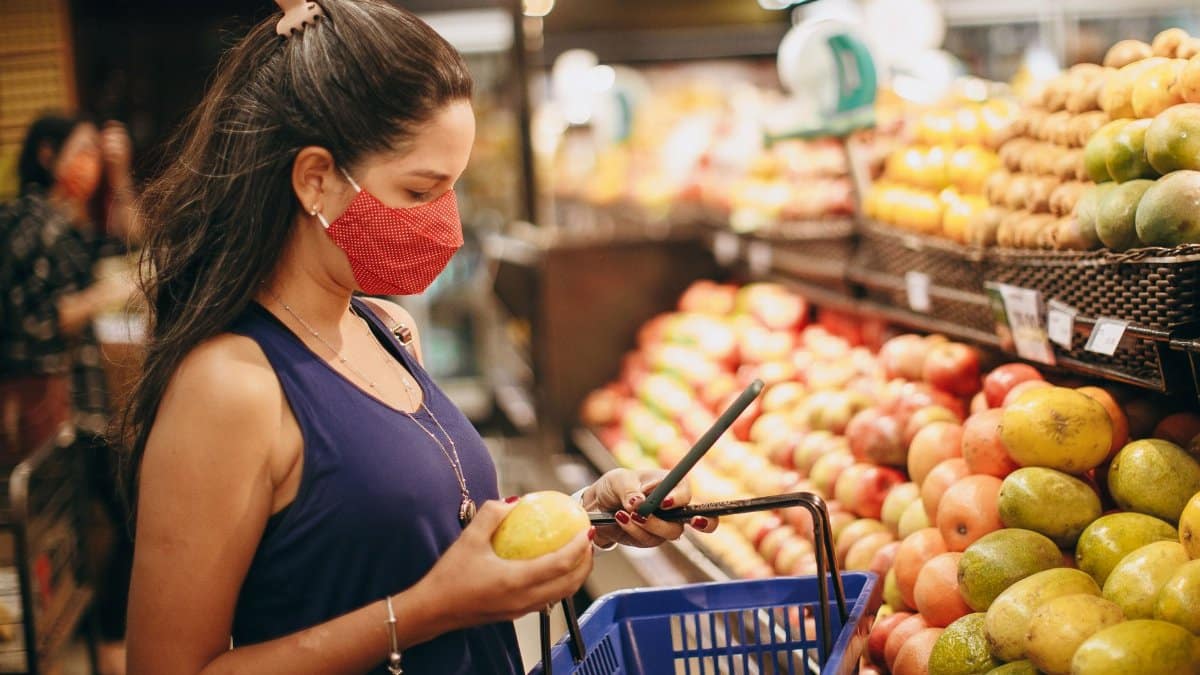 A woman wearing a mask selects fresh fruits in a supermarket produce aisle.