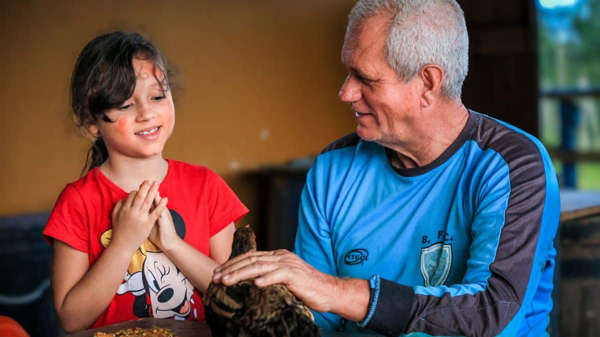 Senior man and young girl interacting with a chicken indoors, fostering bonding and learning.
