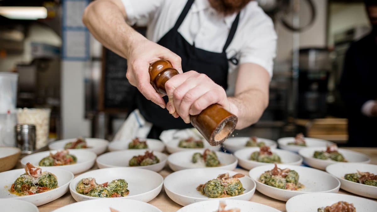 Bearded chef seasoning gourmet dishes with pepper in a busy restaurant kitchen.